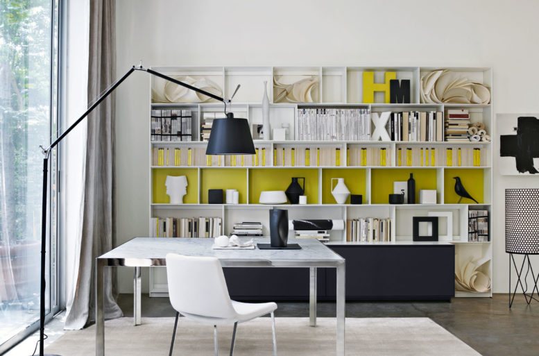 Presenta elementi strutturali in alluminio e piani orizzontali in legno laccato insieme a un sistema di canaline per il passaggio dei cavi.