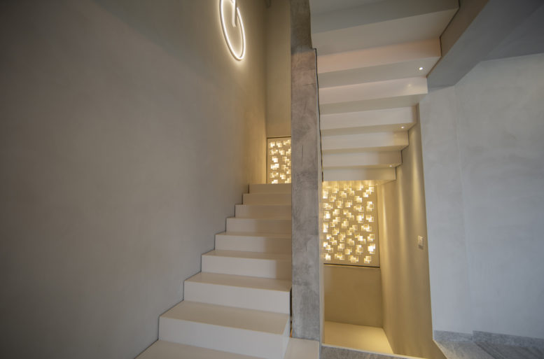 Behind the very fine leather sofas, the white-painted iron staircase seems to float in space, aided by the luminous metal wall. This creates a play of light and shadows. The staircase is double sided and also visible from the entrance and decorates the space.